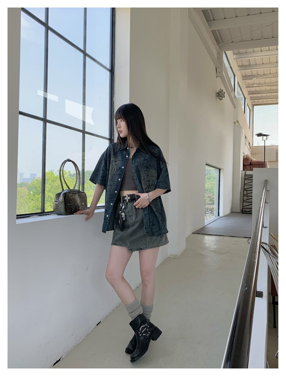 Model wearing the leaf-print short-sleeve shirt open over a crop top with a denim skirt, standing beside a large window