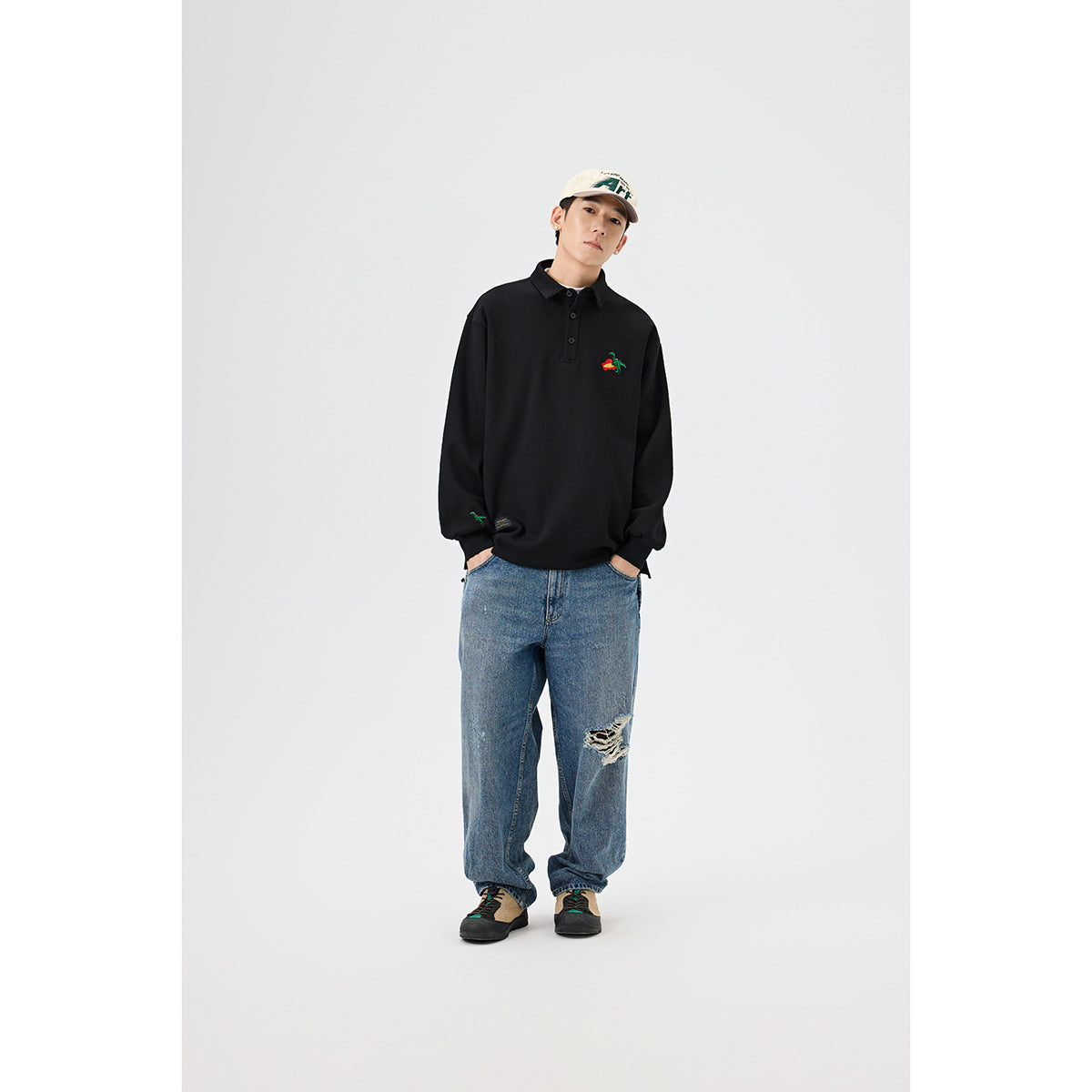 Full-length view of a model wearing a black embroidered long-sleeve collared pullover, loose blue jeans, and a light cap.