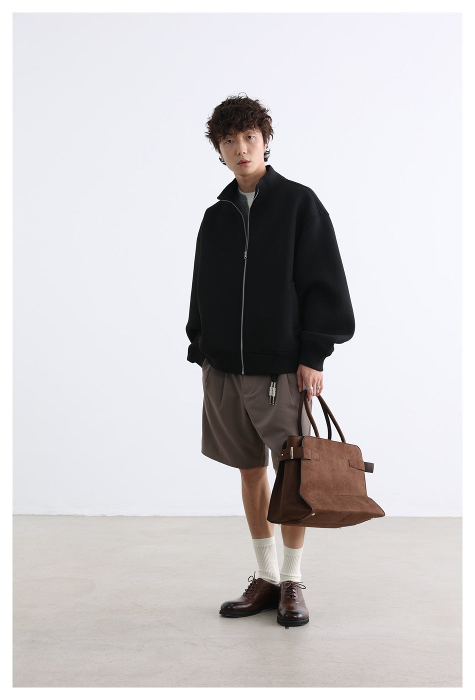 Model wearing a black zip-front jacket with a stand collar and taupe shorts, shown full length from the front.