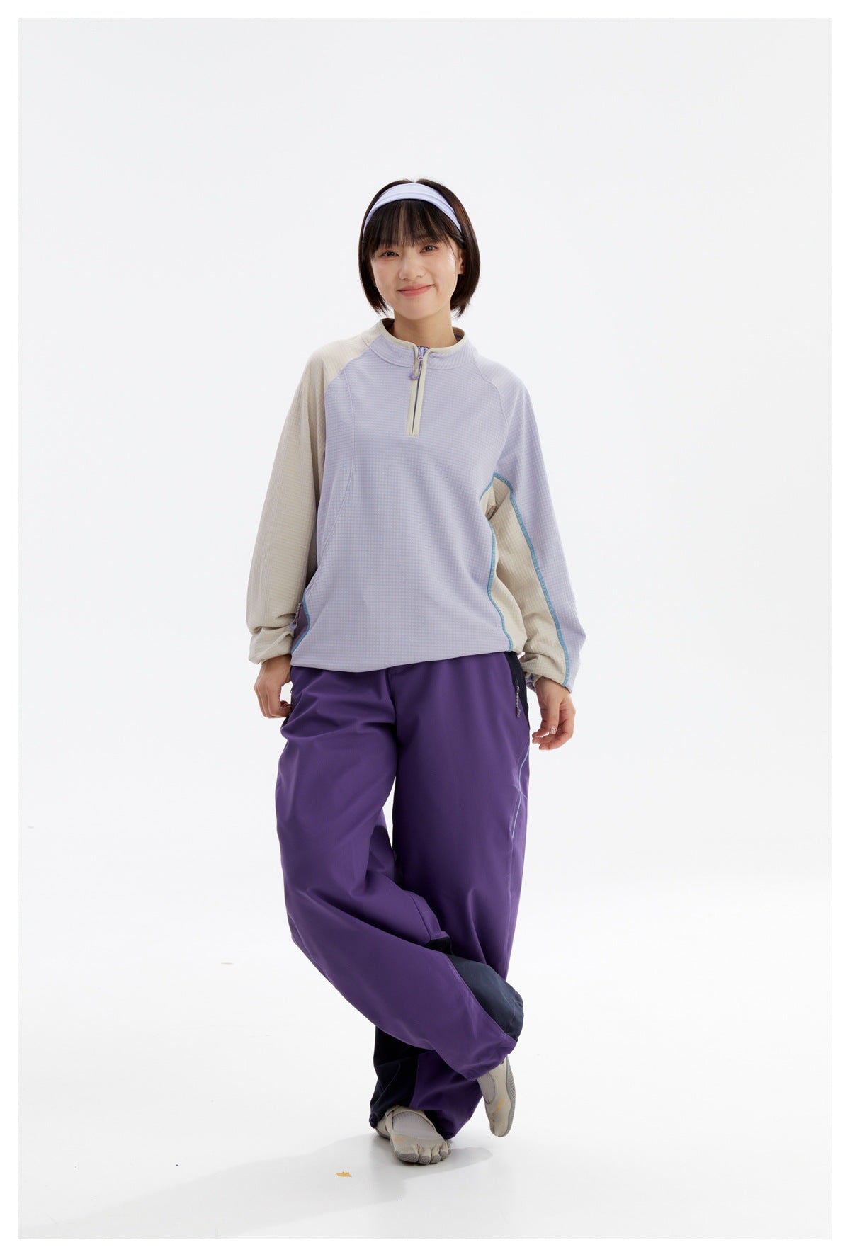 Model wearing a light lavender quarter-zip top with beige sleeves and loose purple pants, standing with crossed legs.