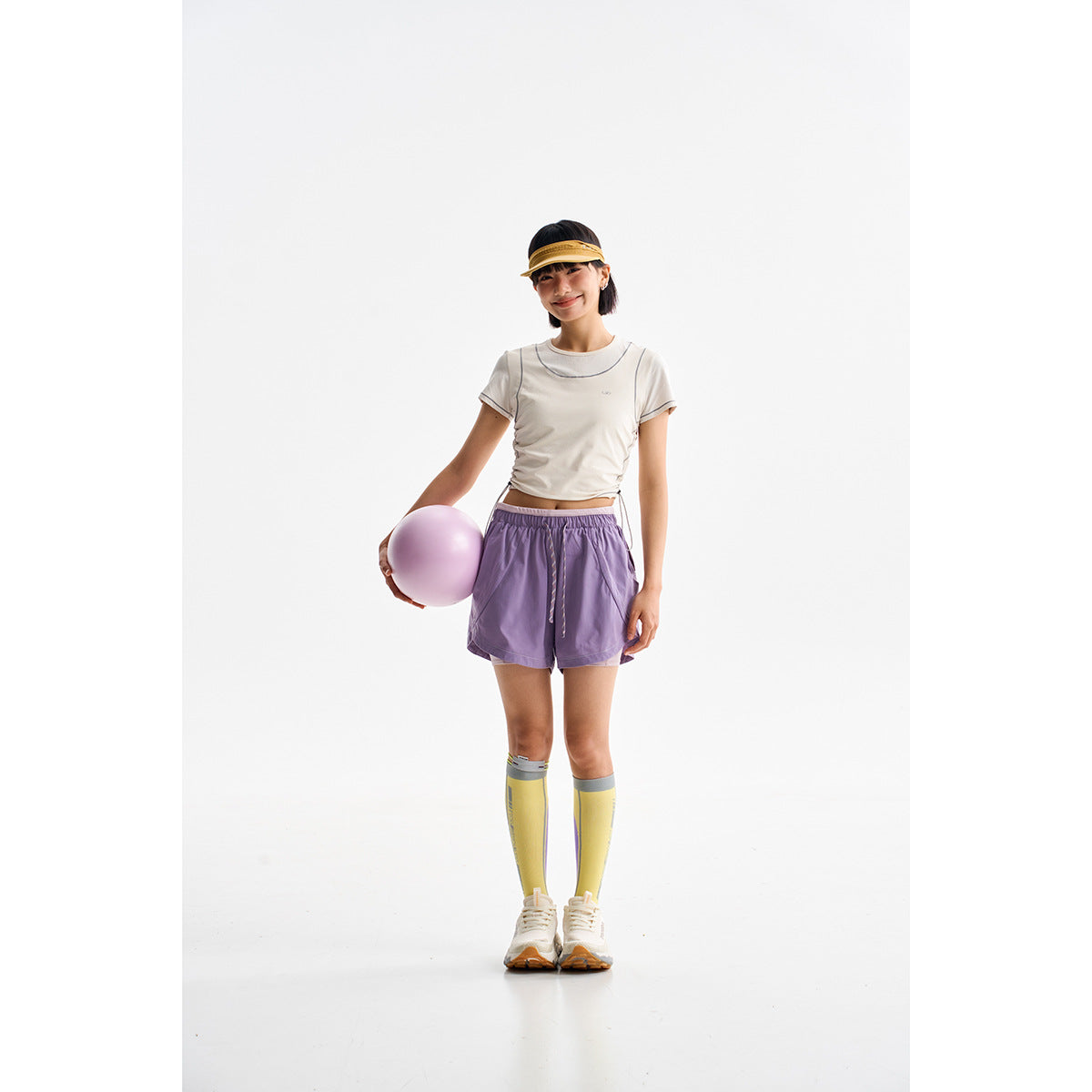 Model wearing an ivory contrast-stitched cropped tee, lavender shorts, yellow knee-high socks and sneakers.