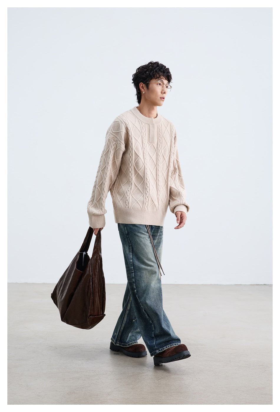 Model wearing a cream cable-knit sweater with diamond panels, loose blue jeans and brown shoes, carrying a dark tote.