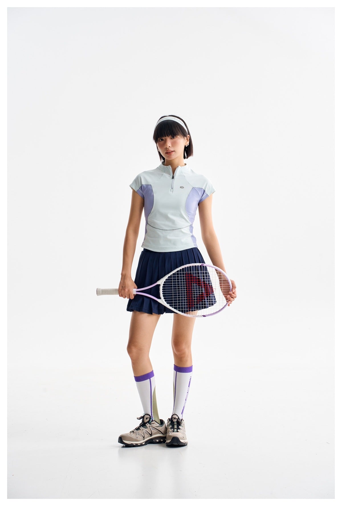 Full-length view of a woman wearing a pale blue quarter-zip top with lavender panels and a navy pleated tennis skirt.