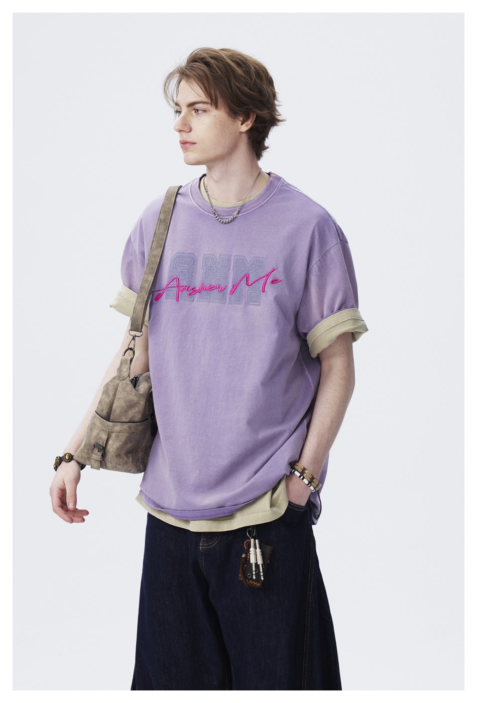 Close three-quarter view of a model in a purple ANM Answer Me T-shirt layered over a khaki tee, with a shoulder bag