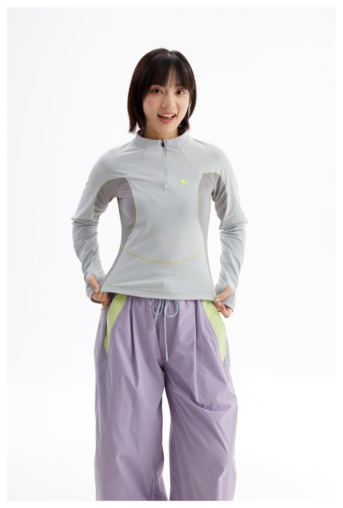 Model wearing a pale gray quarter-zip long-sleeve top with contrasting panels and lime piping, paired with loose lavender