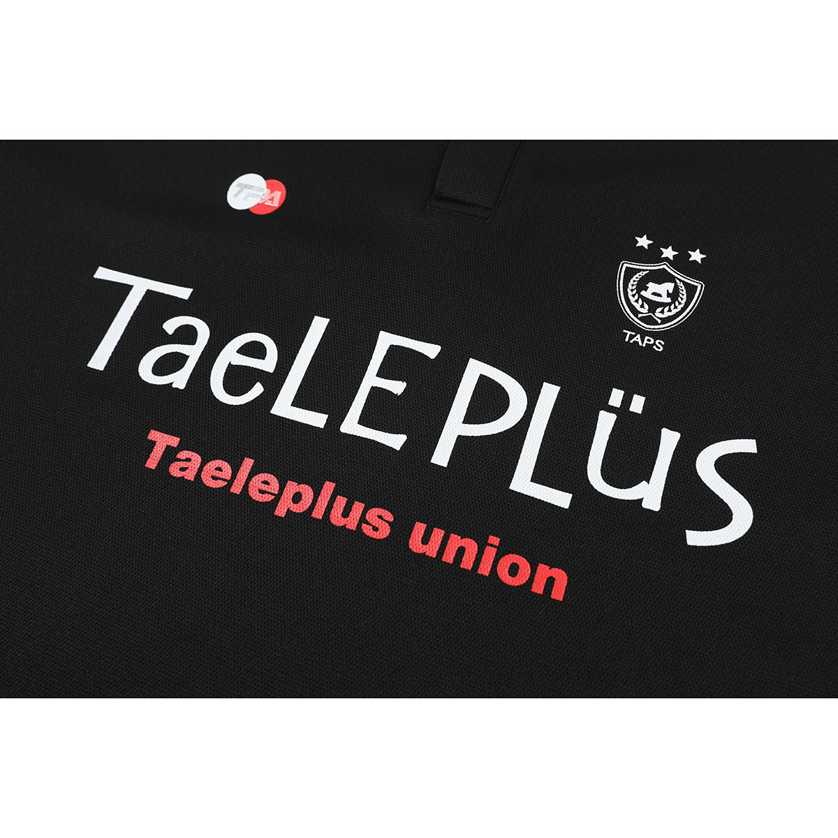 Close-up of a black polo front with large white TaELEPLüs lettering, red text, and a small white crest.