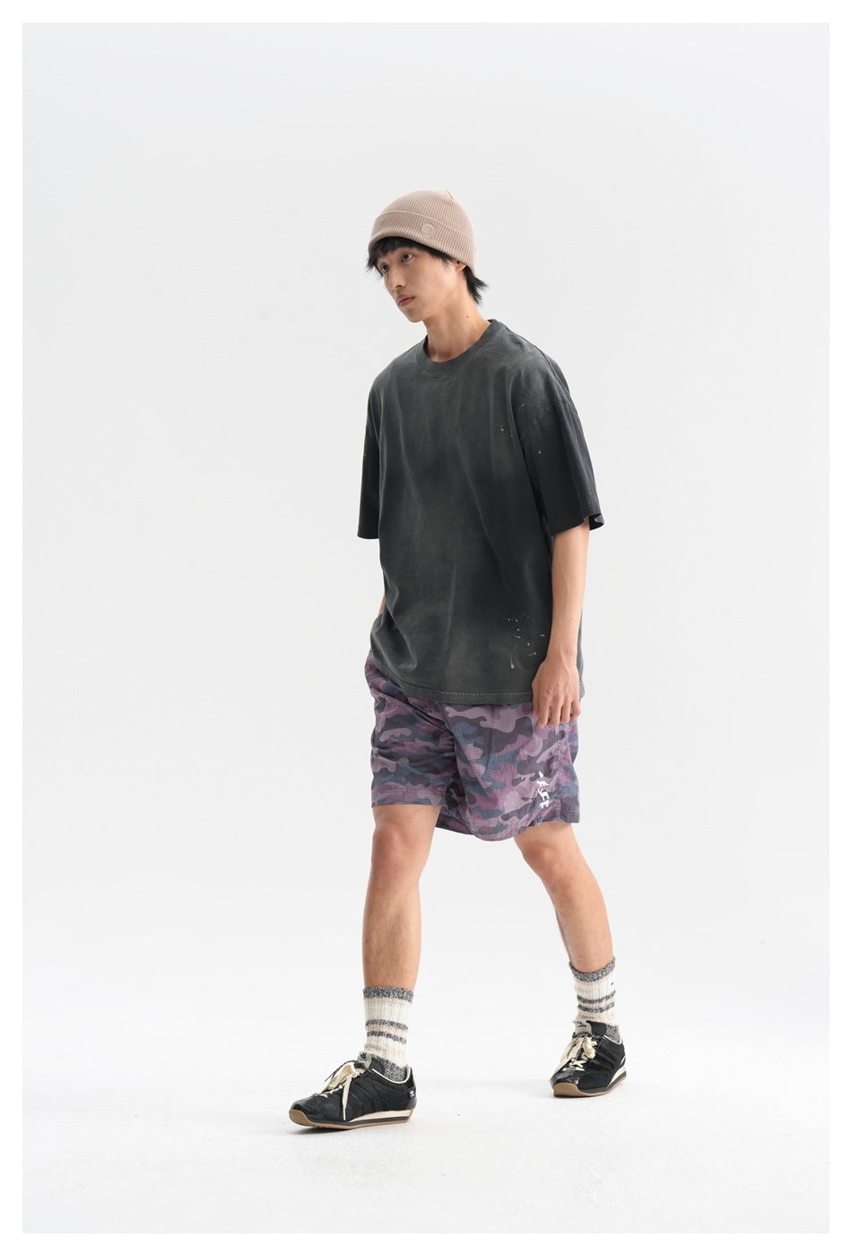 Full-body view of a model wearing an oversized charcoal T-shirt, purple camouflage shorts, and a beige beanie.