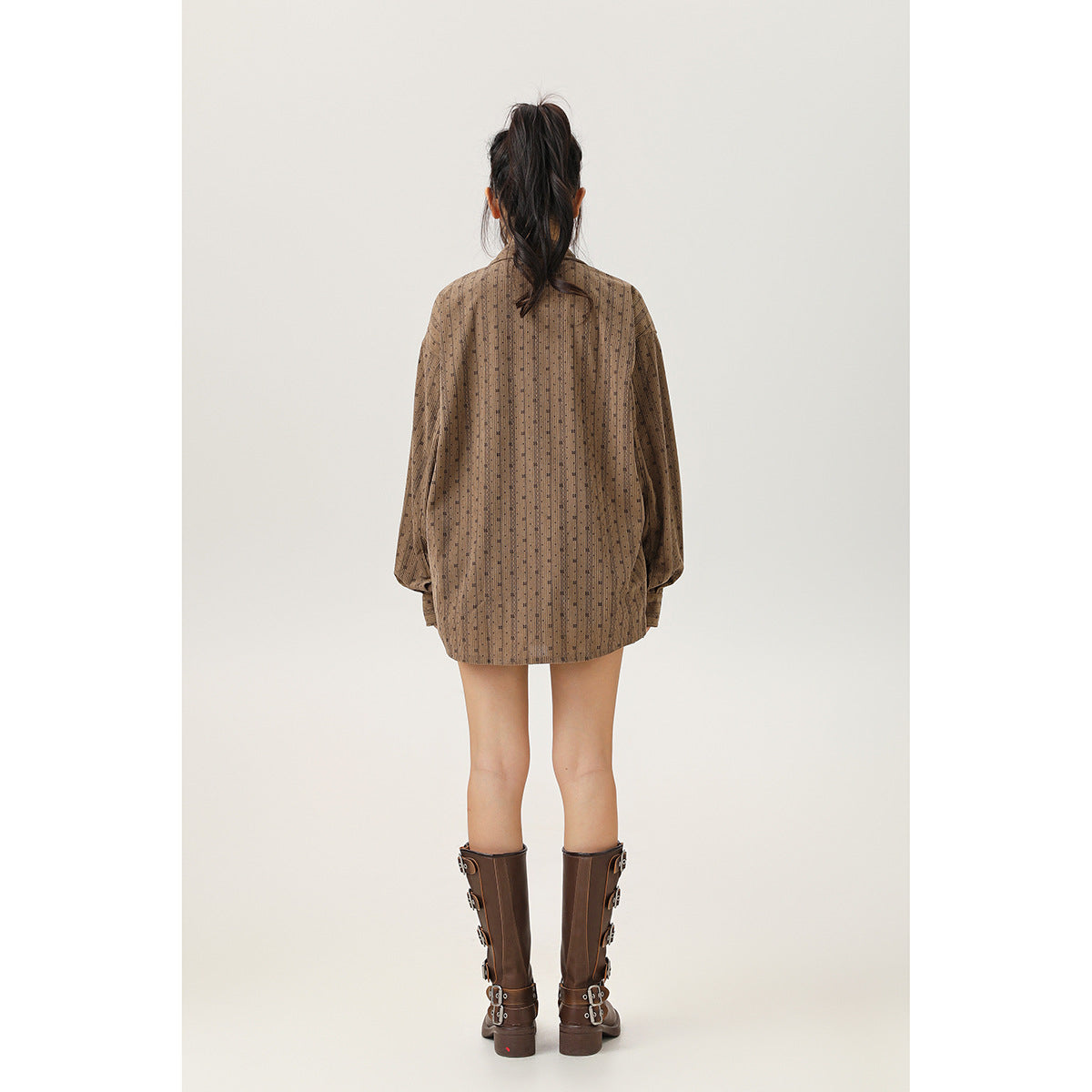Back view of a model wearing an oversized brown shirt with a fine vertical pattern and tall brown boots.