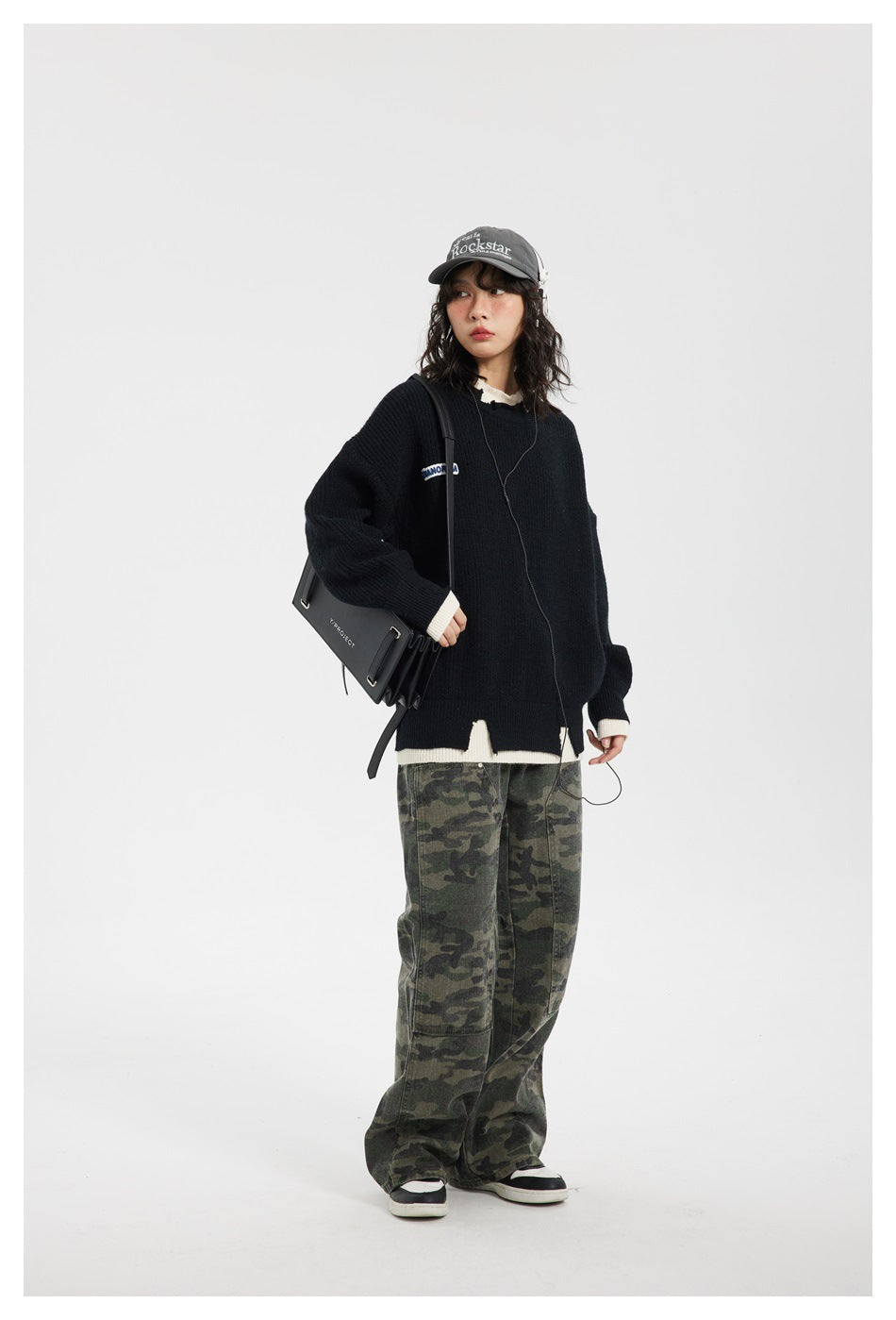 Full-length model wearing an oversized black sweater with cream trim, camouflage pants, and a gray cap.