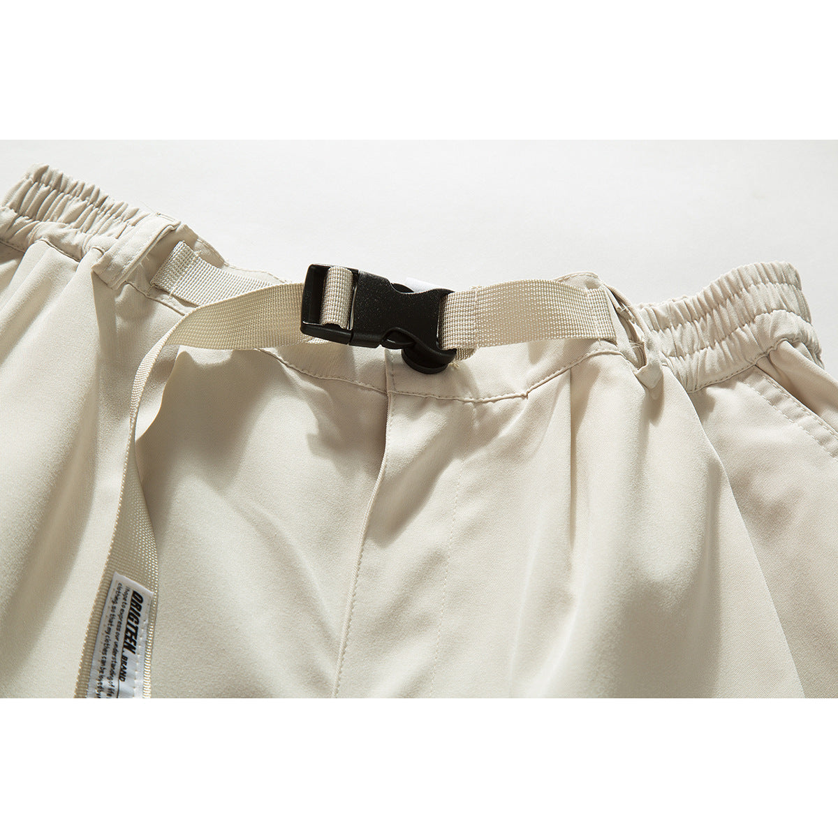 Close-up of cream shorts showing an elastic waistband, adjustable webbing belt with black buckle, and a small sewn label.