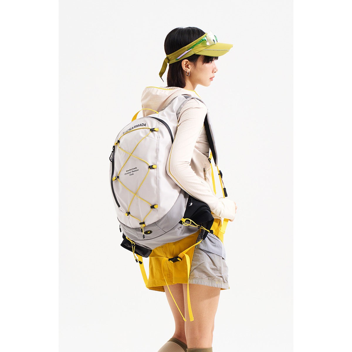 Model in profile wearing a light gray hiking backpack with yellow cord details, a yellow visor, and outdoor clothing.