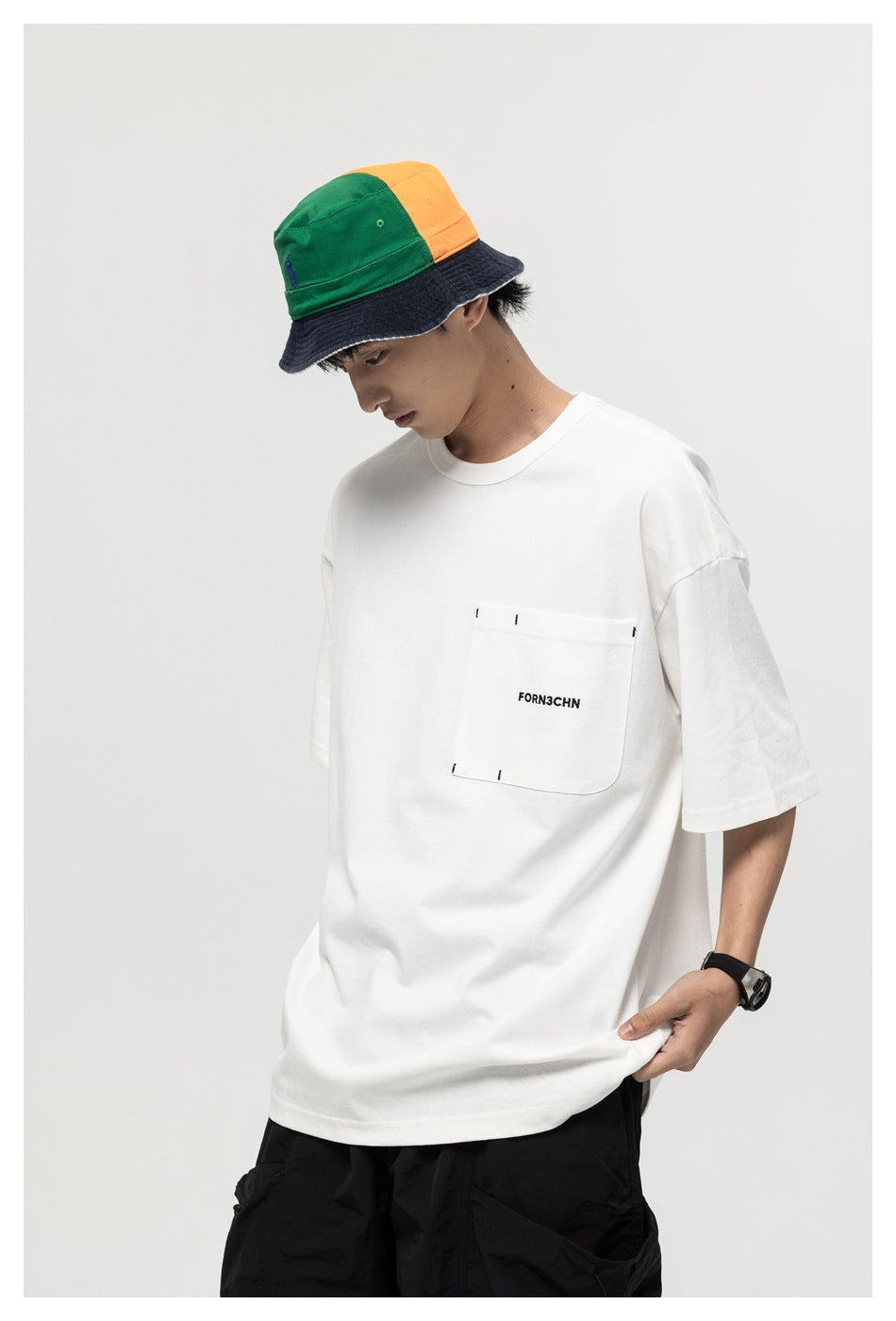 Model wearing an oversized white T-shirt with a chest pocket and small black text, paired with a green, orange, and navy