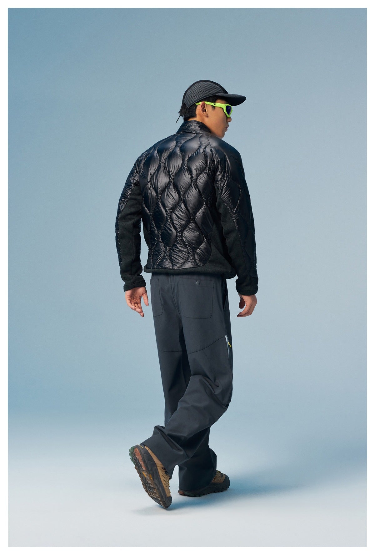 Rear view of a man wearing a black quilted jacket with dark trousers, cap, and neon green sunglasses.