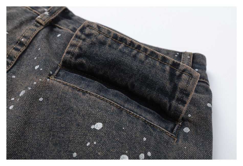 Close-up of a flap pocket on dark washed denim with tan stitching and pale paint splatters.