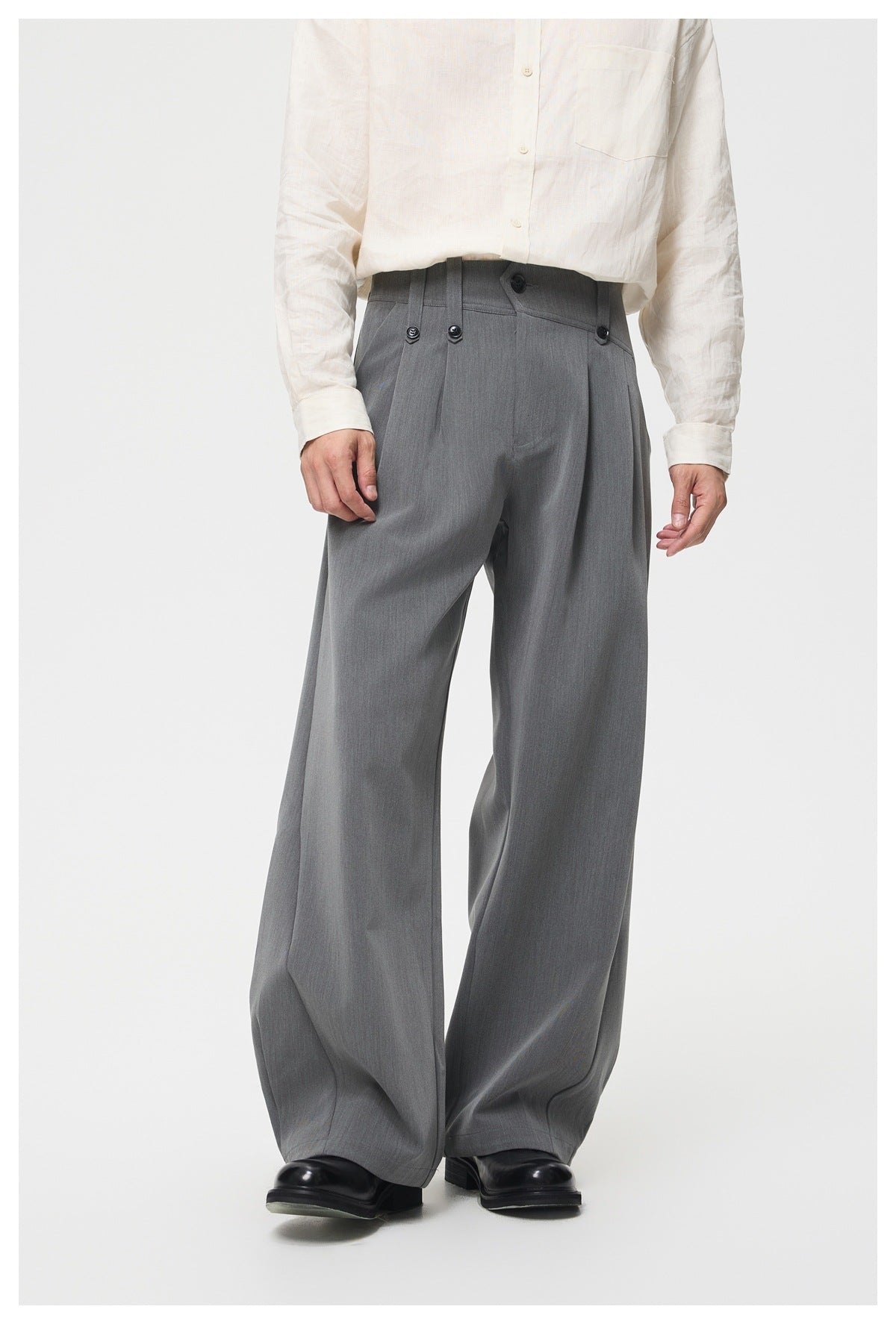 Model wearing gray wide-leg trousers with a buttoned high waist and pleats, styled with an ivory shirt and black shoes.