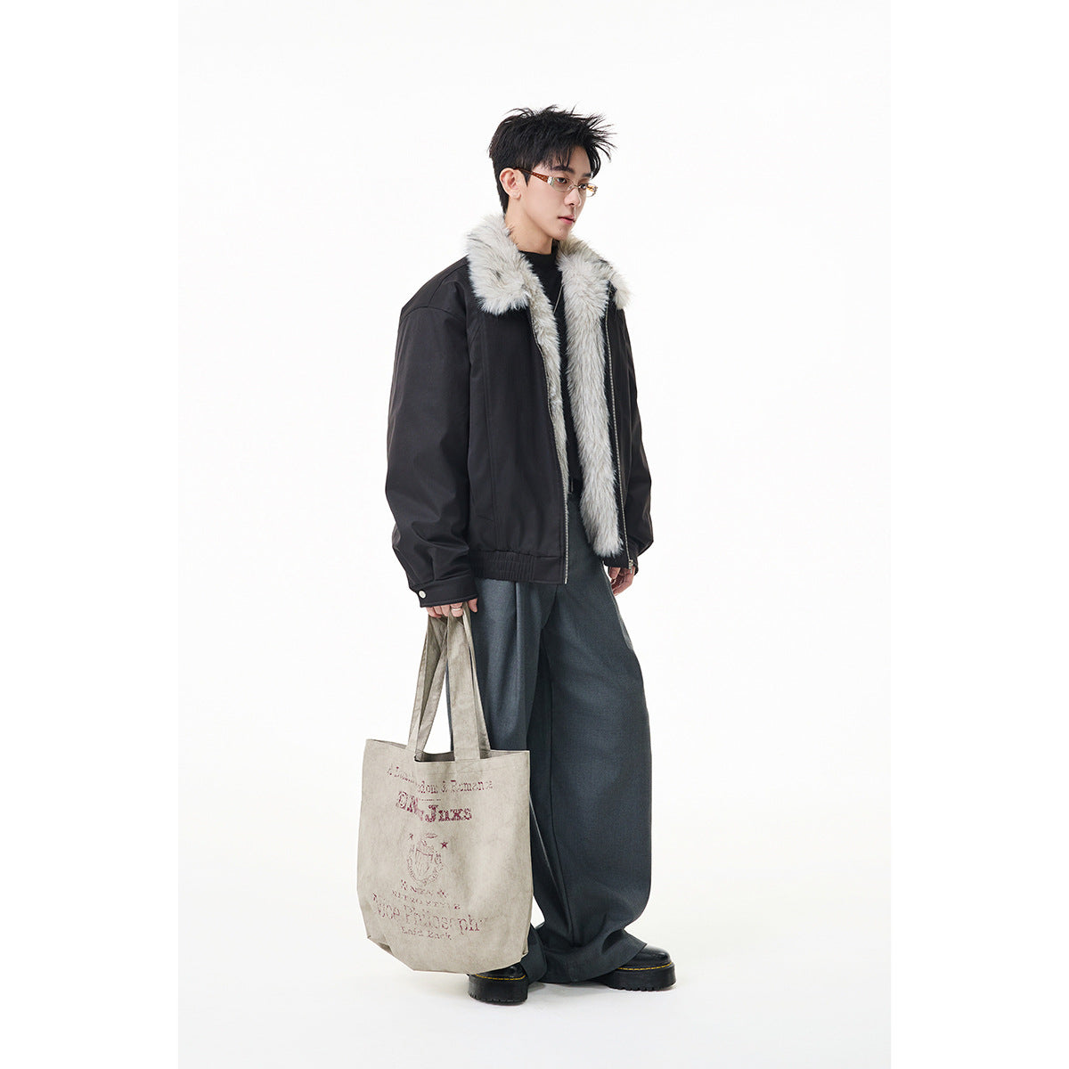 Model wearing a black jacket with a light gray faux-fur collar and lining, wide dark trousers, and carrying a printed tote.