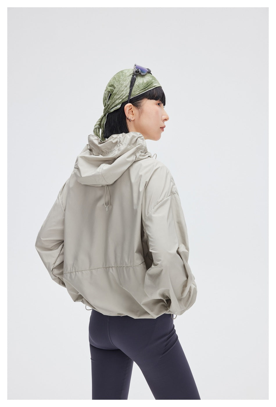 Back view of a model wearing a light gray hooded jacket with a gathered hem and navy leggings.