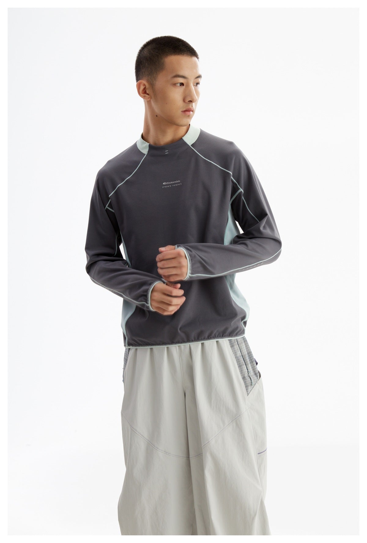 Model wearing a charcoal long-sleeve top with pale mint accents and loose light gray pants, adjusting one sleeve.