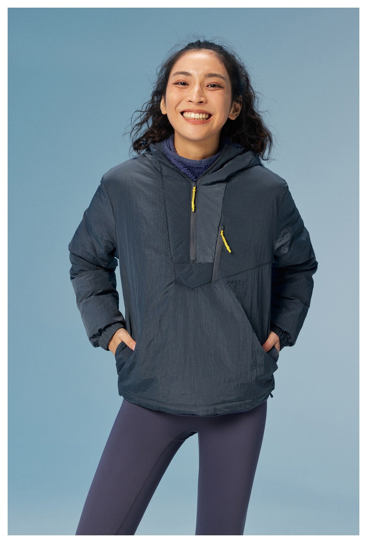 Front view of a woman wearing a dark slate hooded pullover with yellow zipper pulls and purple leggings.