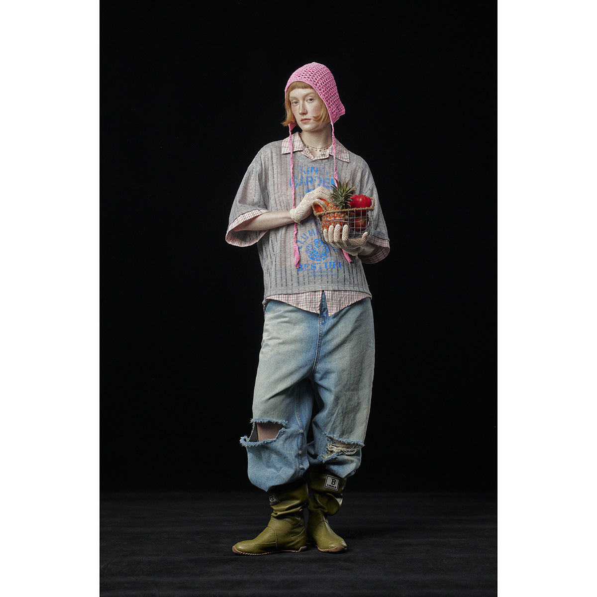 Model wearing a gray blue-graphic tee over a checked shirt, distressed jeans, a pink crochet bonnet, and olive boots.