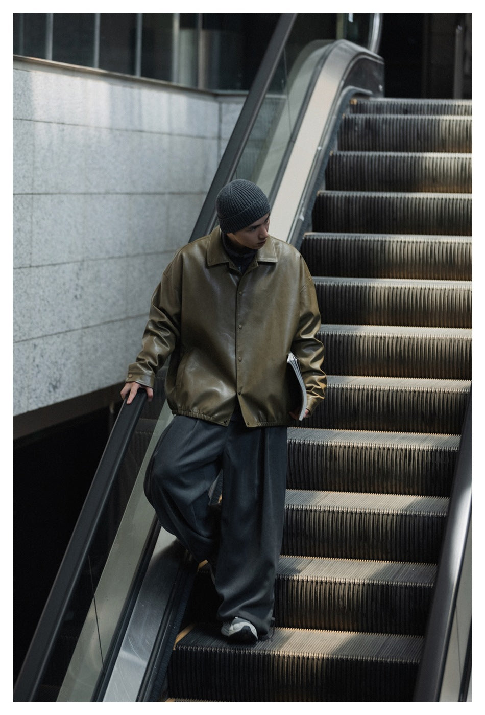 Model in a glossy olive-brown button-front jacket, charcoal wide-leg pants, and a dark ribbed beanie on an escalator.