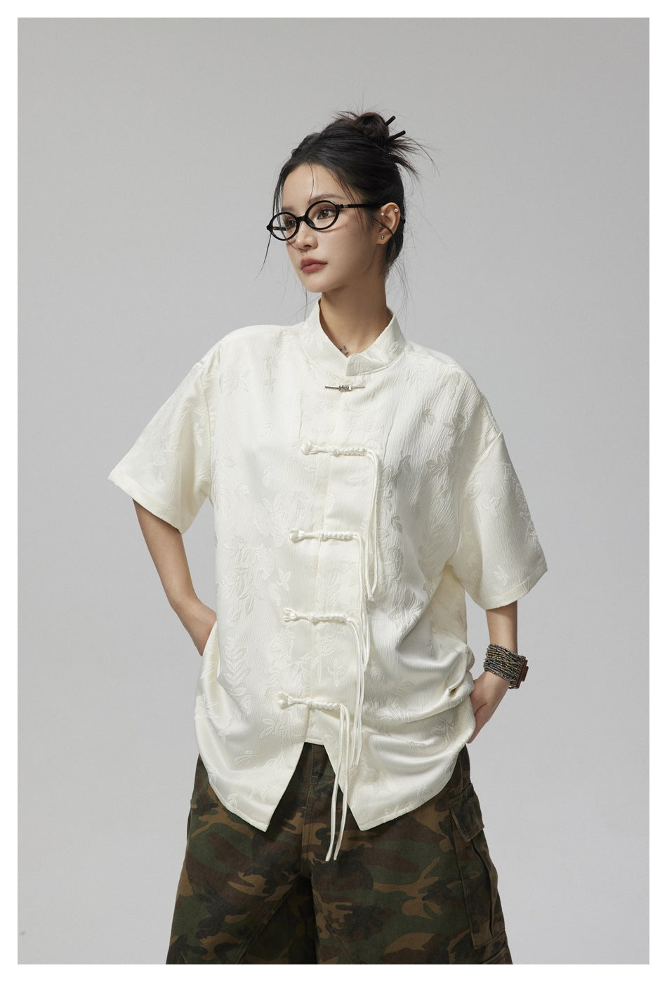 Model wearing an ivory floral short-sleeve shirt with a stand collar and braided closures, paired with camouflage trousers.