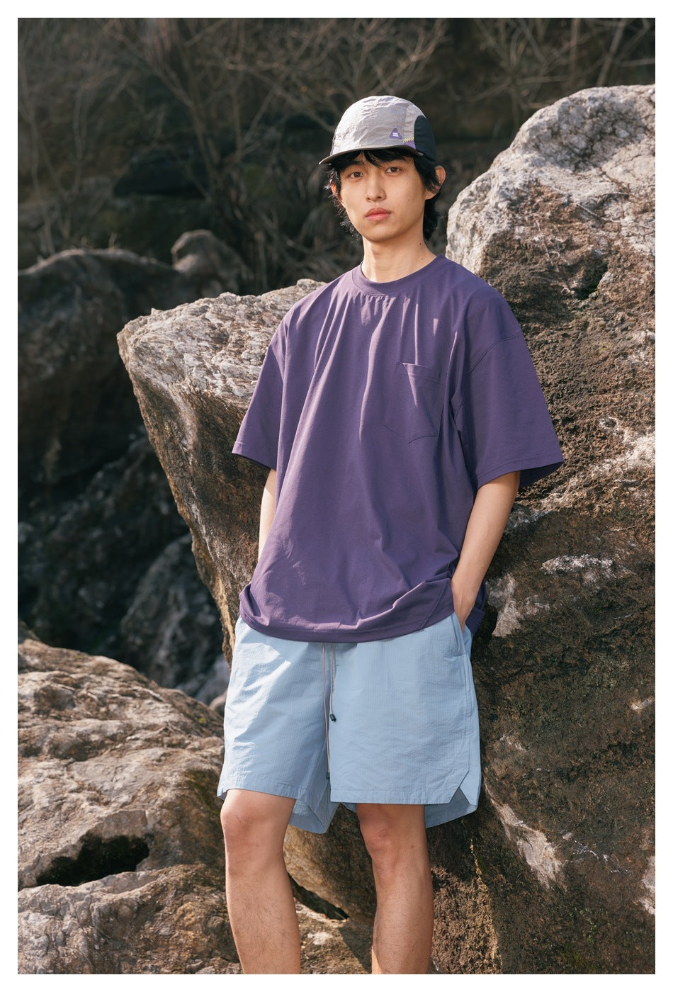 Front view of a model wearing a purple pocket T-shirt and light blue shorts among rocks.