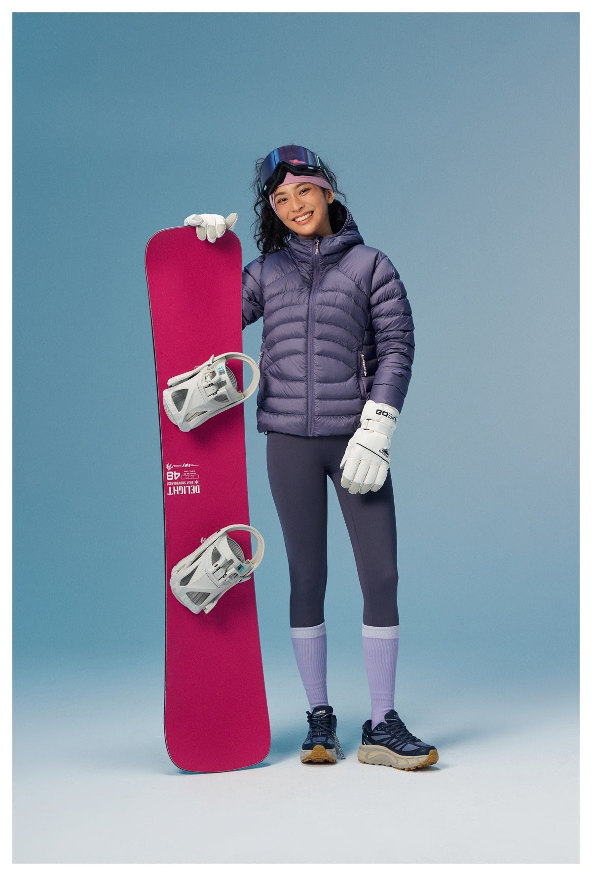 Woman wearing a purple hooded quilted puffer jacket with leggings, holding a snowboard, front view