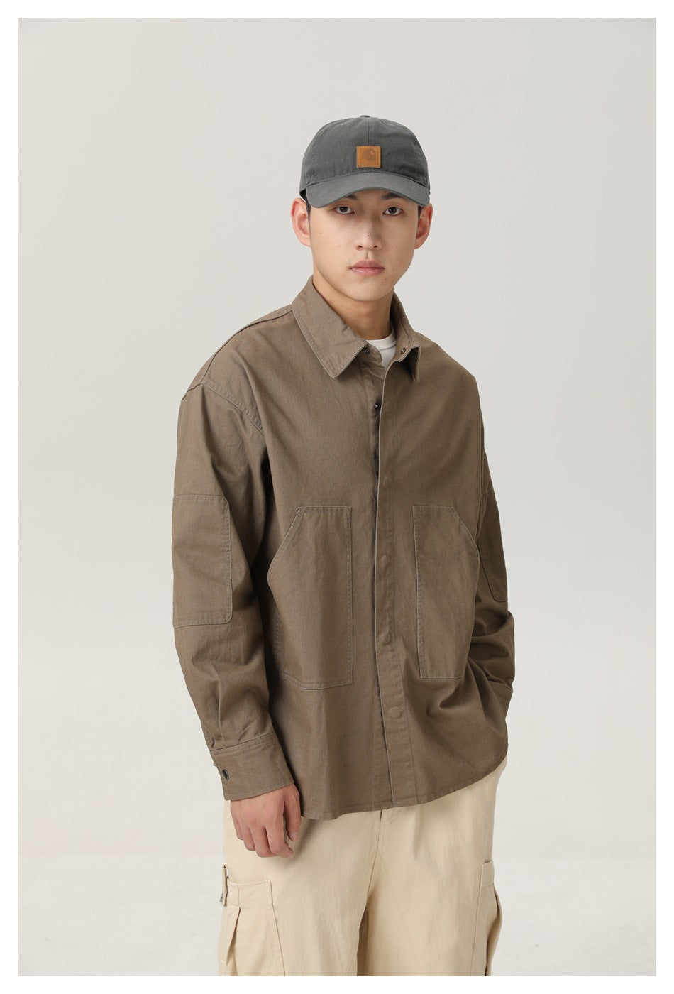 Model in a brown button-front overshirt with large front and sleeve pockets, cream cargo pants and a dark cap.