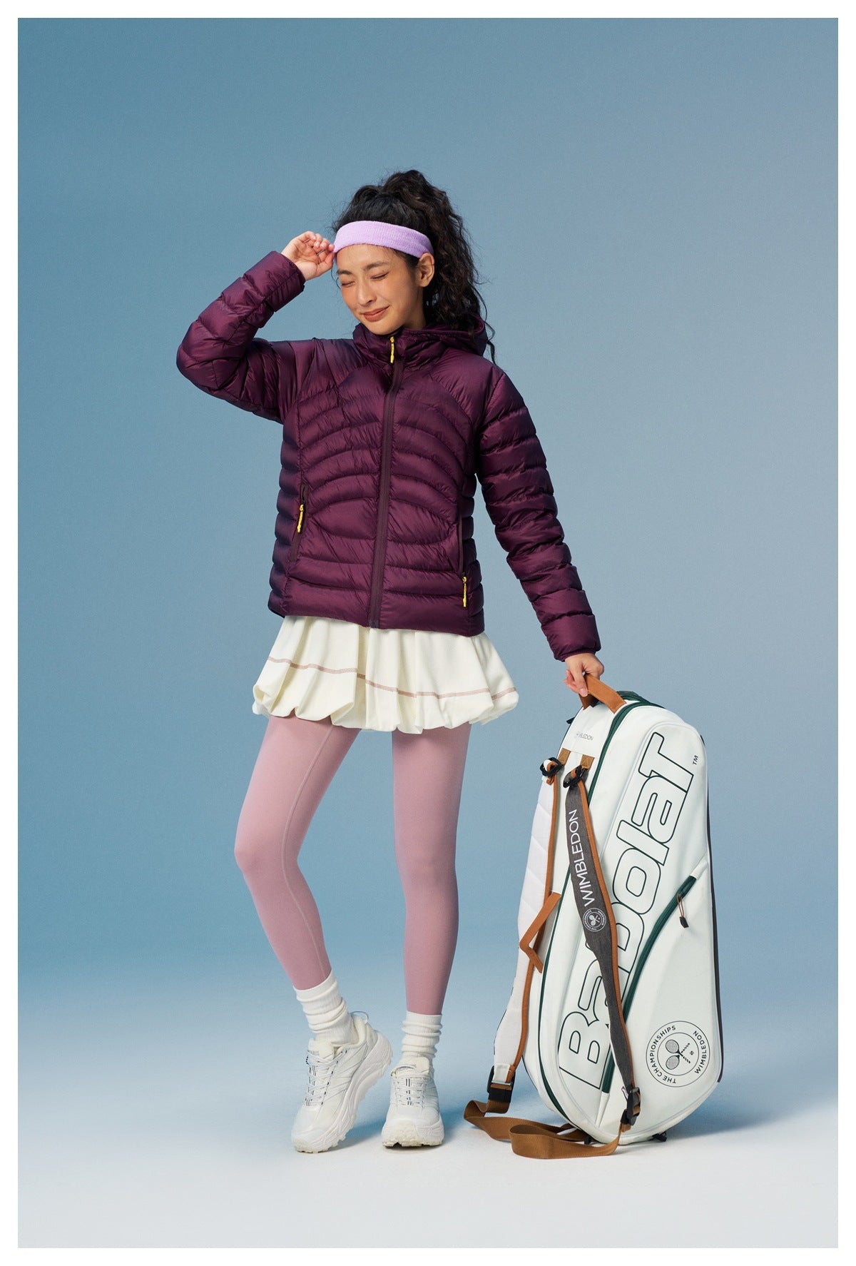 Model wearing a plum quilted hooded jacket with yellow zipper pulls, white tennis skirt, and pink leggings.