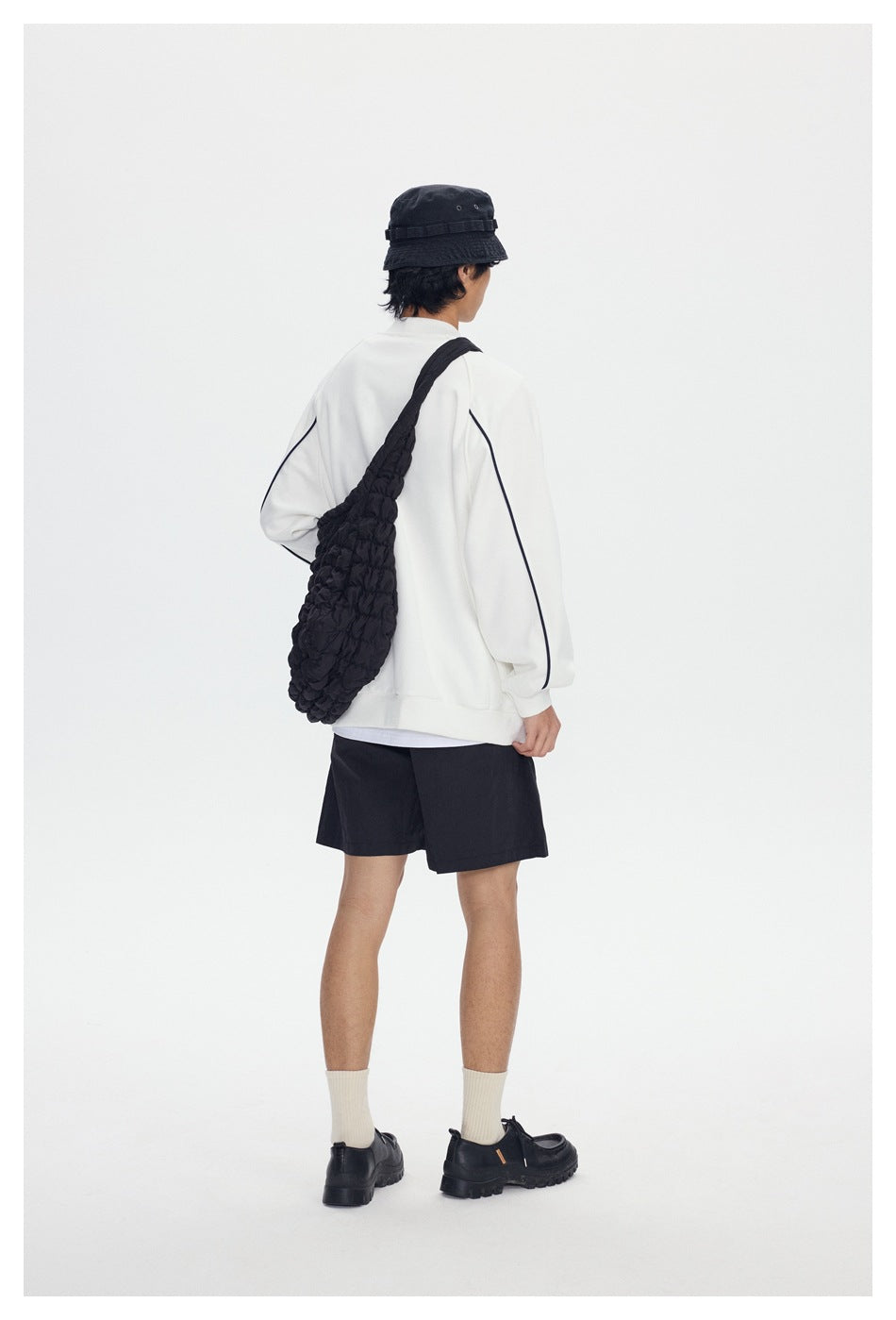 Rear view of a model wearing a white sweatshirt with navy piping and black shorts, carrying a black shoulder bag.
