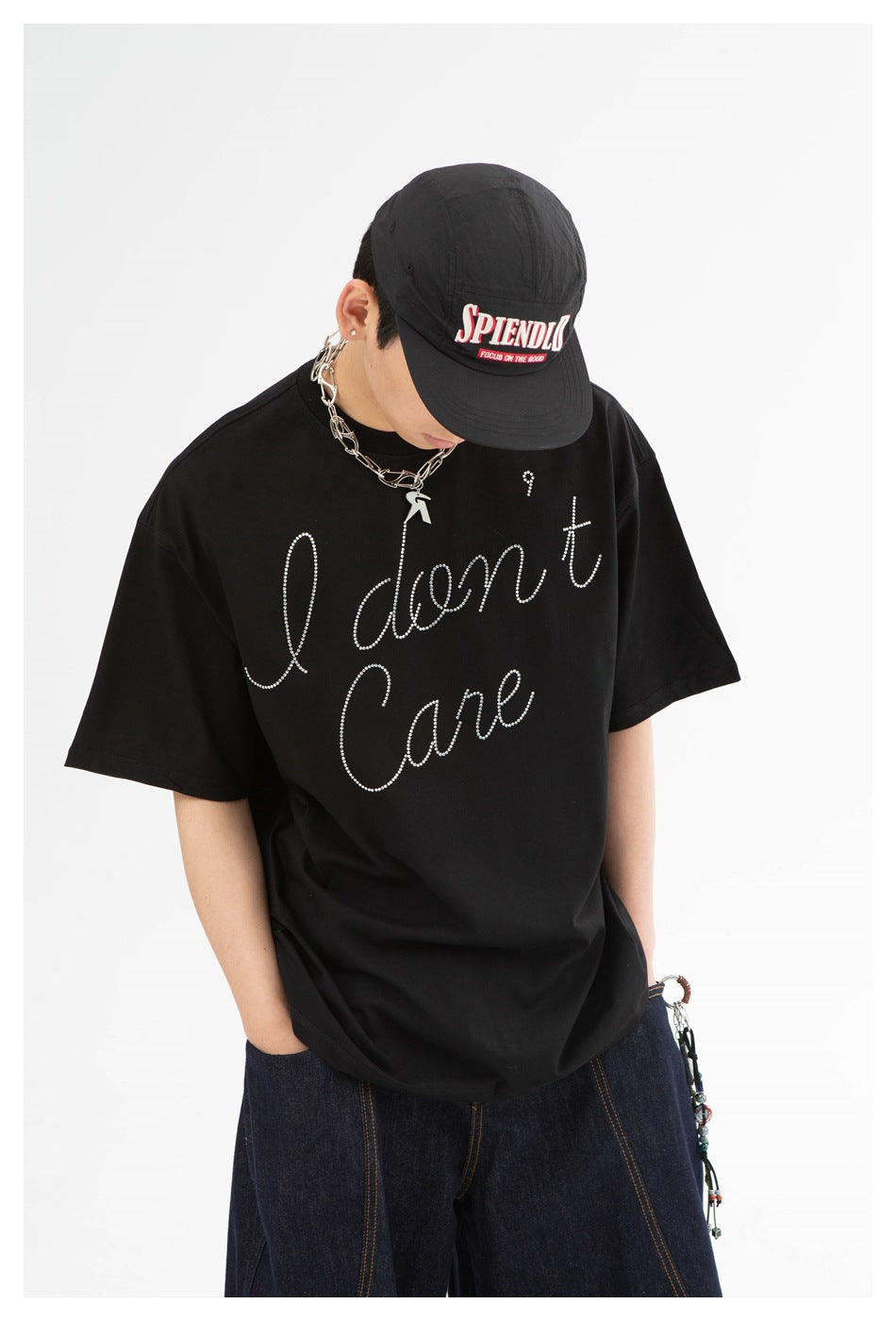 Model looking down in a black oversized rhinestone-script T-shirt and black cap, with dark denim bottoms.