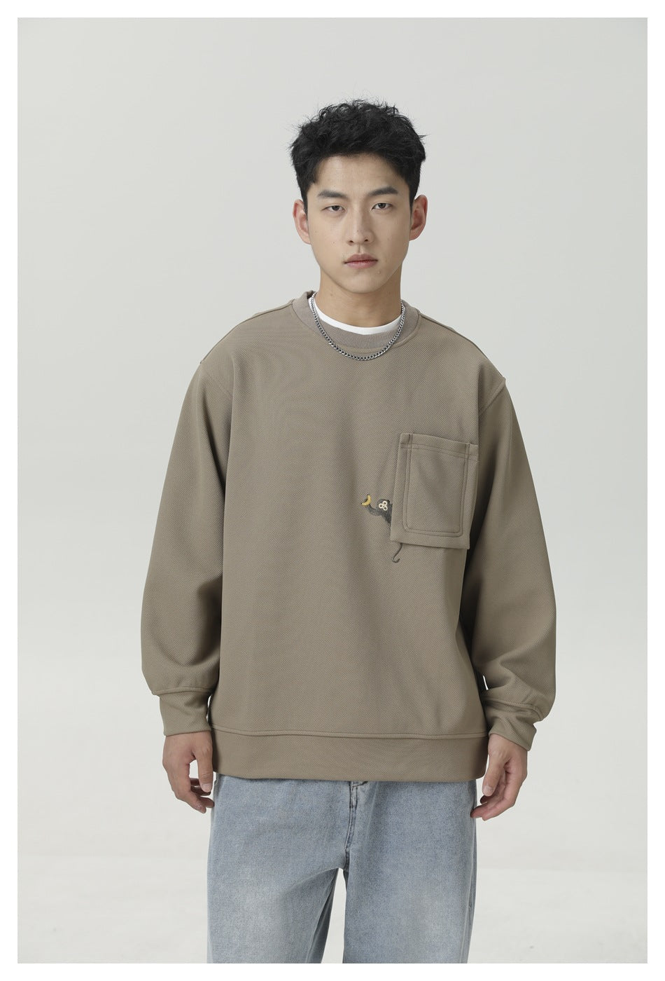 Front view of a man wearing an oversized taupe crewneck sweatshirt with a chest pocket and light blue jeans.