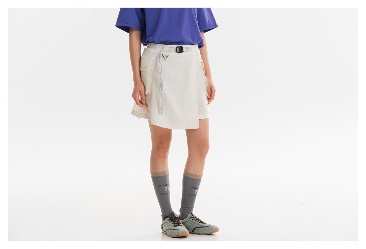 Front view of a model wearing an ivory belted wrap skort with utility pockets, a purple T-shirt, gray socks and sneakers.