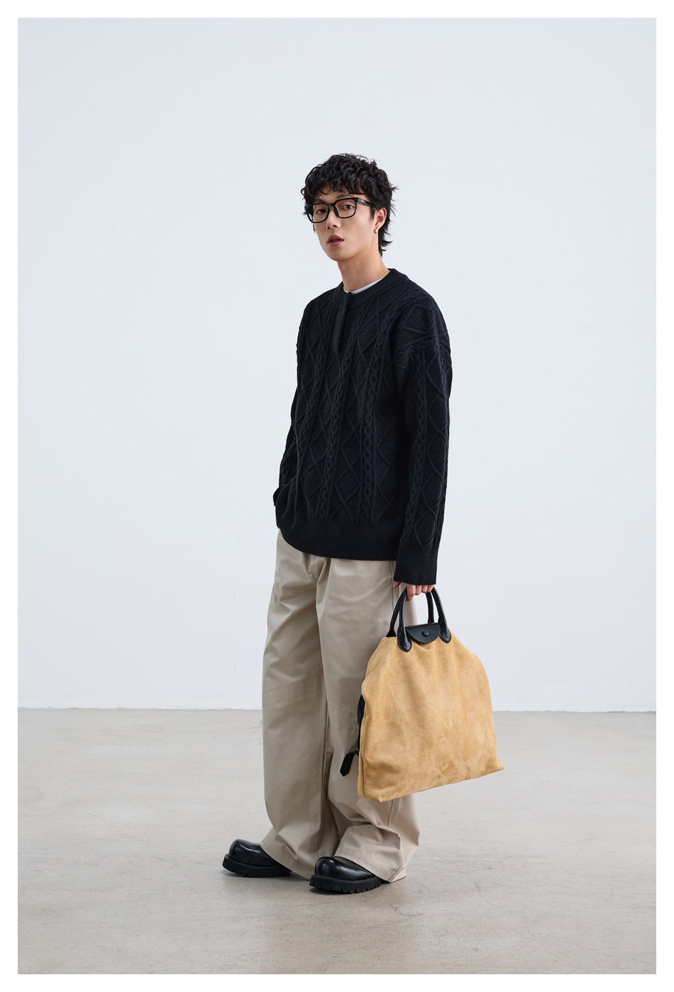 Model wearing a black diamond-pattern cable-knit sweater with beige trousers and carrying a tan bag.