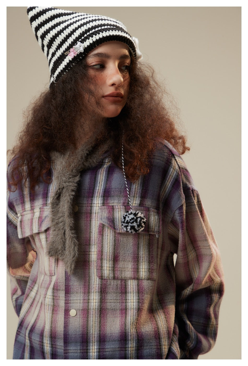 Close-up of a model wearing a purple and pink plaid jacket with flap pockets, gray fuzzy trim, and a striped knit hat.