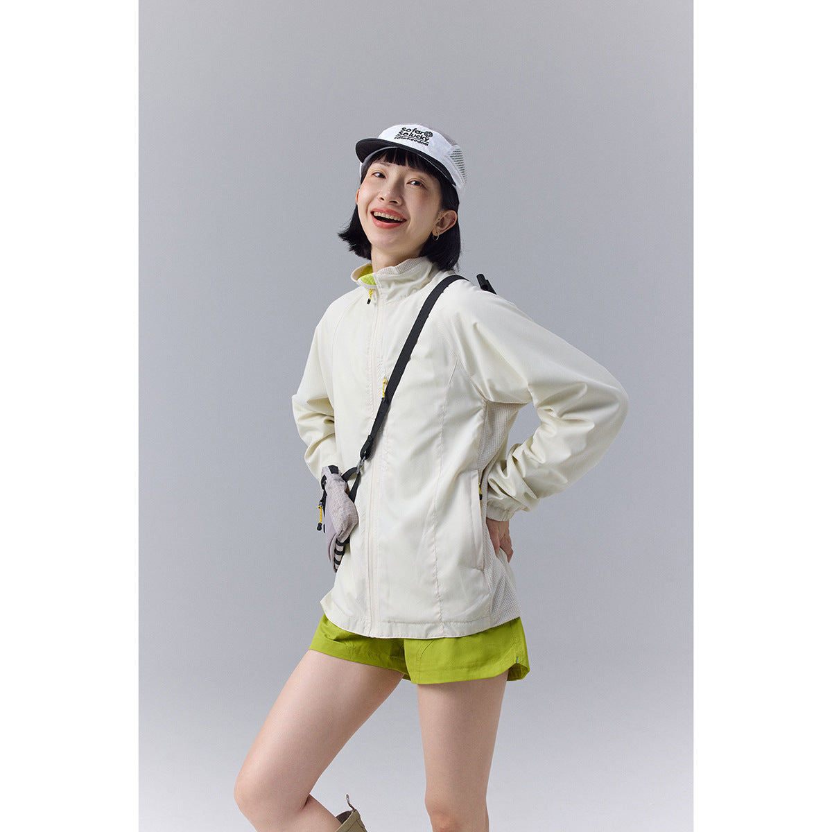 Model wearing a cream zip-front jacket with lime accents and bright green shorts, shown with a crossbody bag.