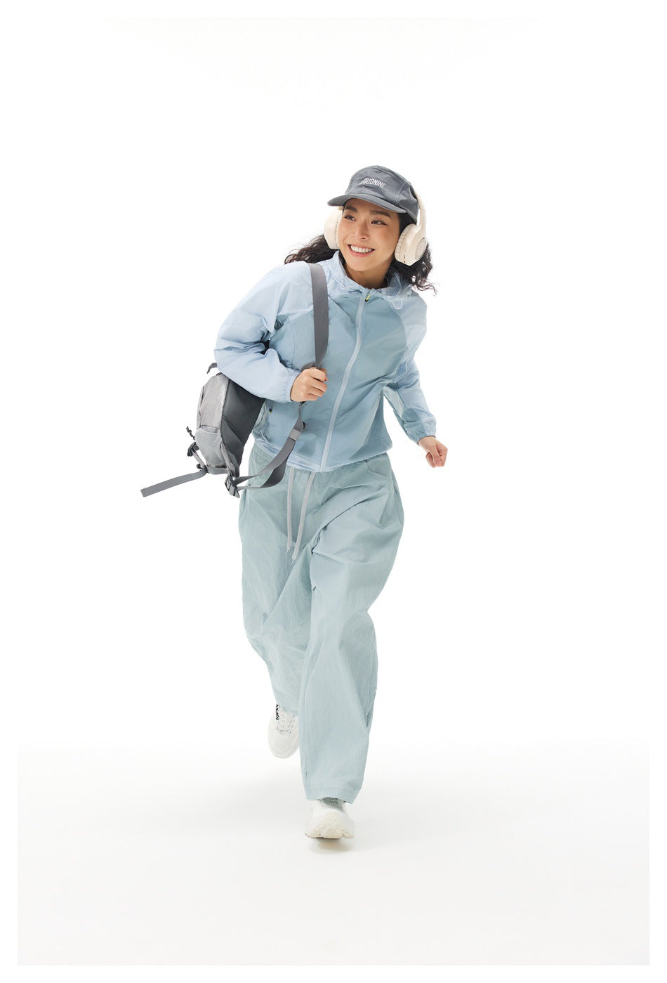 Model in a light blue hooded jacket and loose pale blue pants, running with a gray backpack.