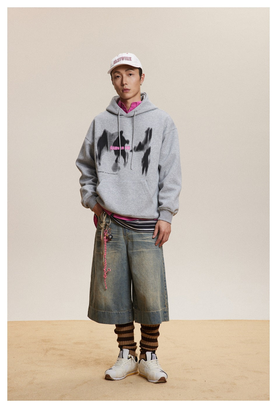 Full-length model wearing a gray graphic hoodie, white cap, denim shorts and striped leg warmers.