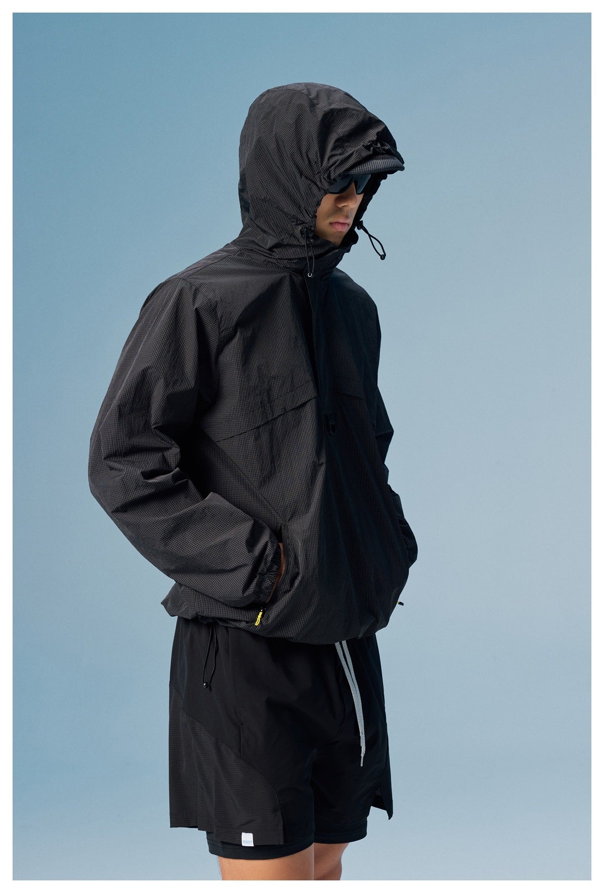 Side view of a man wearing a black grid-textured hooded windbreaker and black shorts.