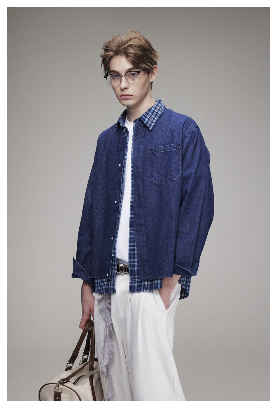 Three-quarter view of a model wearing a navy denim overshirt over a blue plaid shirt and white trousers, with a cream bag.