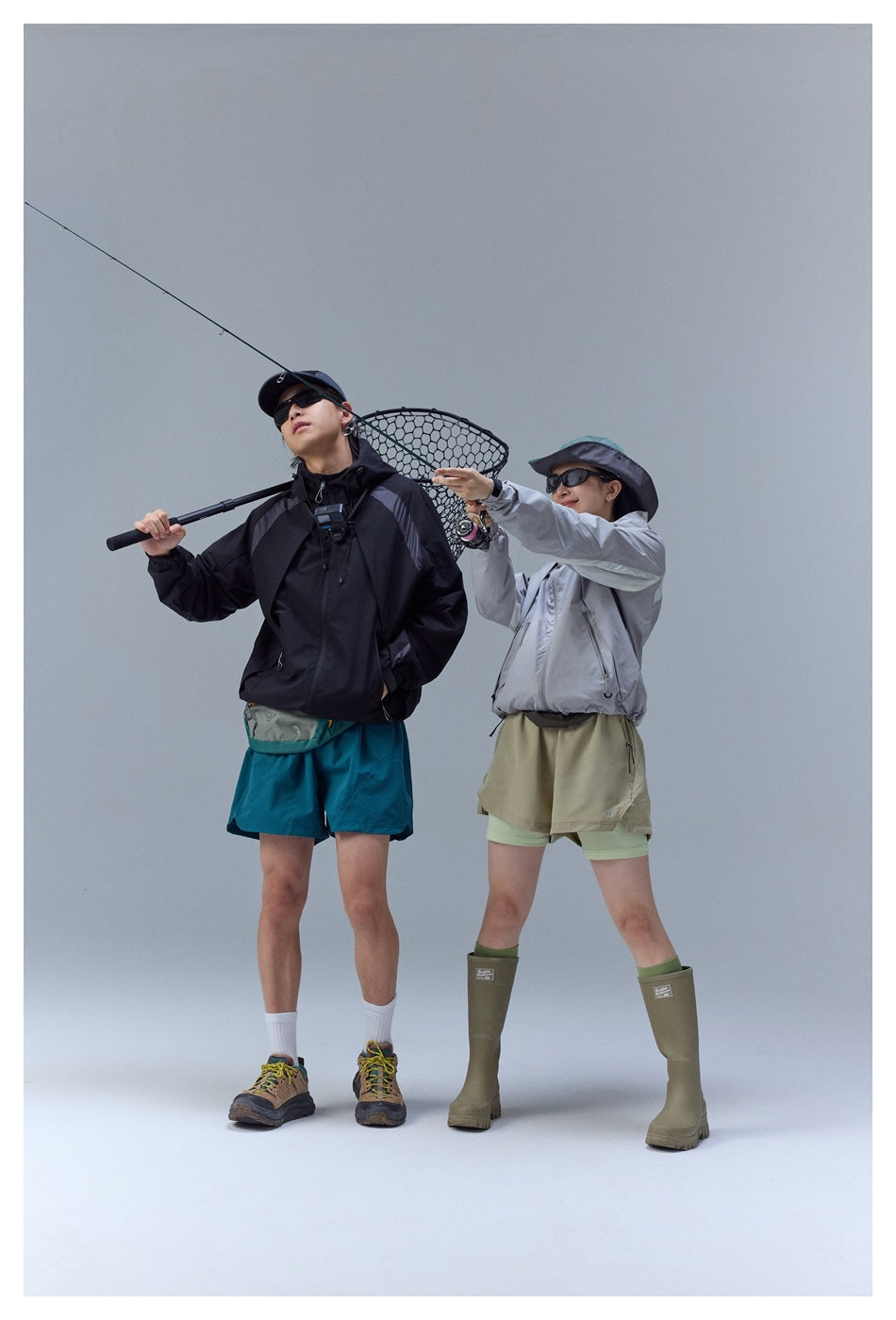 Two models wear black and silver hooded jackets with teal and khaki shorts while posing with fishing gear.