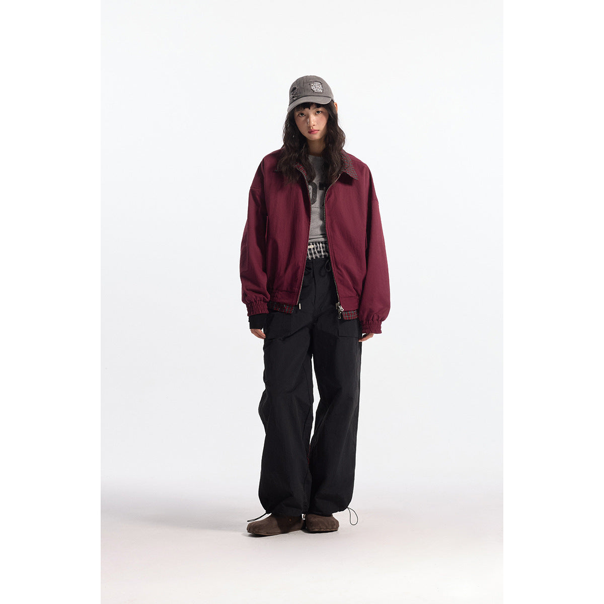 Model wearing an oversized burgundy zip jacket with plaid trim, black wide-leg pants, a gray cap and brown shoes.