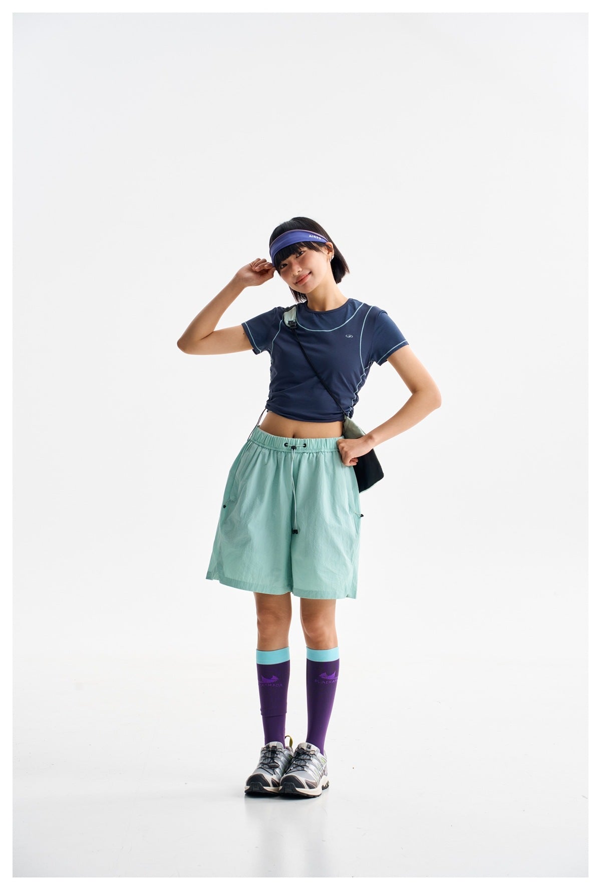 Model in a navy piped tee and mint-green shorts, wearing a purple headband and dark crossbody bag.