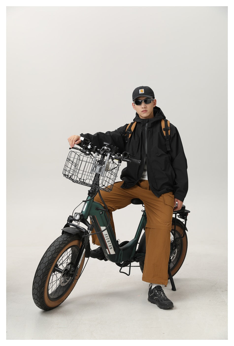 Model wearing a black hooded jacket and brown cargo trousers while seated on a bicycle.