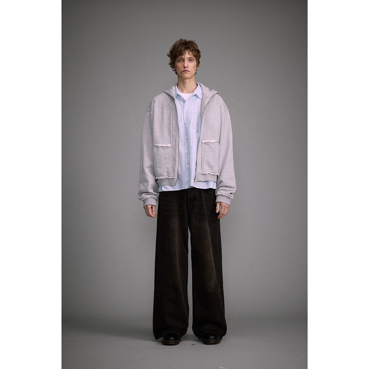 Full-length model wearing a light gray zip hoodie with frayed pocket edges, a blue striped shirt, and wide black trousers.