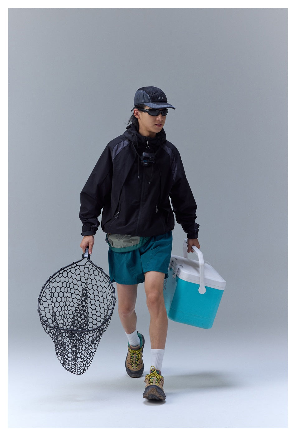 Model wearing a black hooded jacket and teal shorts, carrying a fishing net and a turquoise cooler.