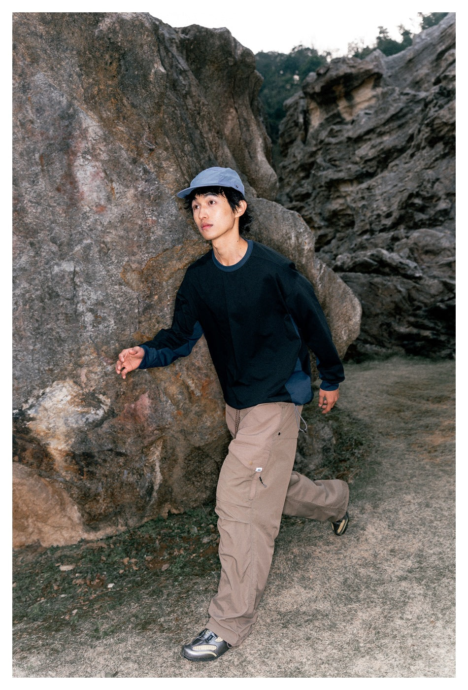 Model wearing a dark long-sleeve shirt and loose taupe cargo pants while walking beside rocks.