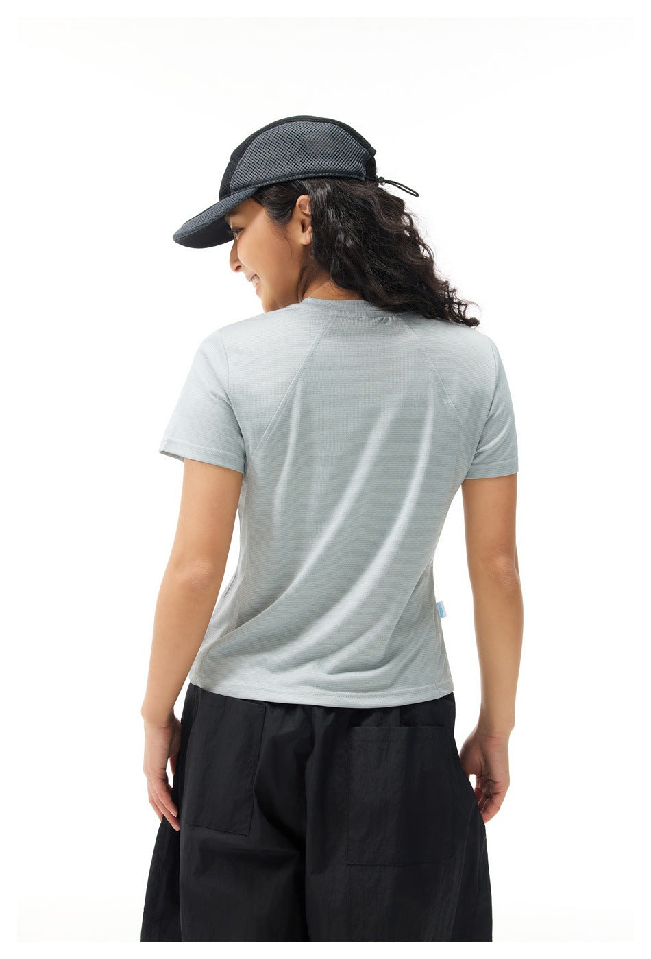 Back view of a model wearing a pale gray short-sleeve shirt with diagonal shoulder seams and loose black pants.