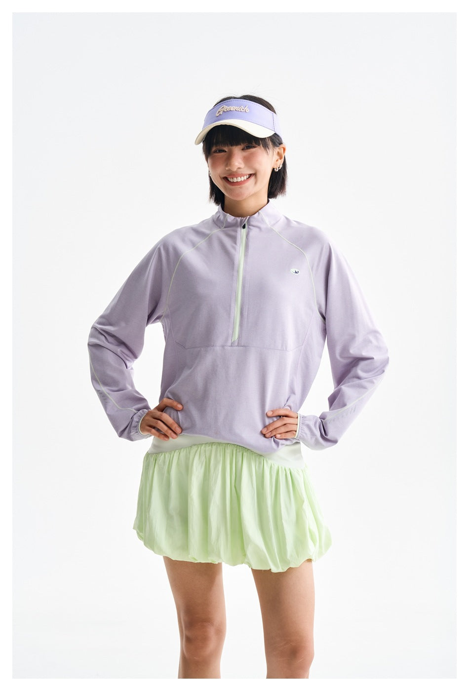 Front view of a smiling model wearing a lilac quarter-zip top, pale mint bubble skirt and lavender visor.